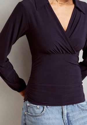 Kobieta w dopasowanym czarnym długim topie z kopertowym dekoltem w serek i jasnoniebieskich dżinsach, ręce opierające się na biodrach.