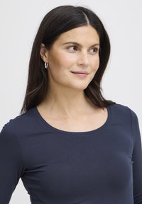 Cămașă bleumarin cu mâneci lungi și decolteu rotund. Textură netedă, croială ajustată. Modelul poartă cercei argintii cerc și are păr drept și închis la culoare.