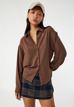 Frau trägt eine beige Kappe, ein lockeres braunes Hemd mit Knöpfen und einen blauen karierten Rock und steht mit einer Hand in der Hüfte vor einem neutralen Hintergrund.
