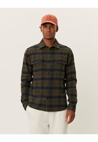 Geruit shirt in olijfgroen en marineblauw, met twee voorzakken, een knoopsluiting en lange mouwen. Vergezeld van een lichtoranje pet.