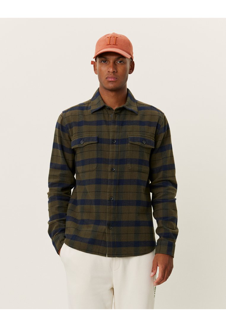 Geruit shirt in olijfgroen en marineblauw, met twee voorzakken, een knoopsluiting en lange mouwen. Vergezeld van een lichtoranje pet.
