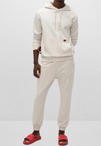 Felpa beige con tasca frontale e logo bianco, abbinata a pantaloni jogger coordinati. Ciabatte rosse con logo sulla cinghia, che mostrano un design semplice.