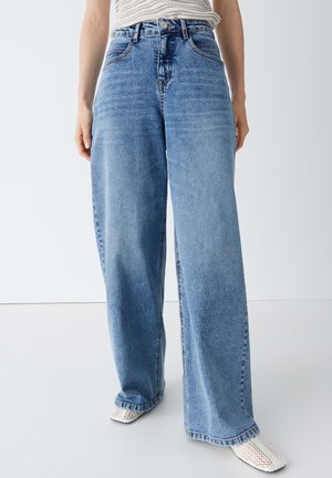 Vrouw die een blauwe high-waist wijd been jeans draagt en witte geweven schoenen met vierkante neus, staand tegen een effen achtergrond.