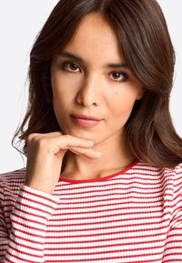 Woman with long, dark hair, wearing a red and white striped ribbed top. She rests her chin on her hand, with a neutral expression.