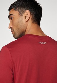 Rød kortermet skjorte med rund hals og en glatt tekstur. Har en hvit "Venum" logo på øvre rygg.