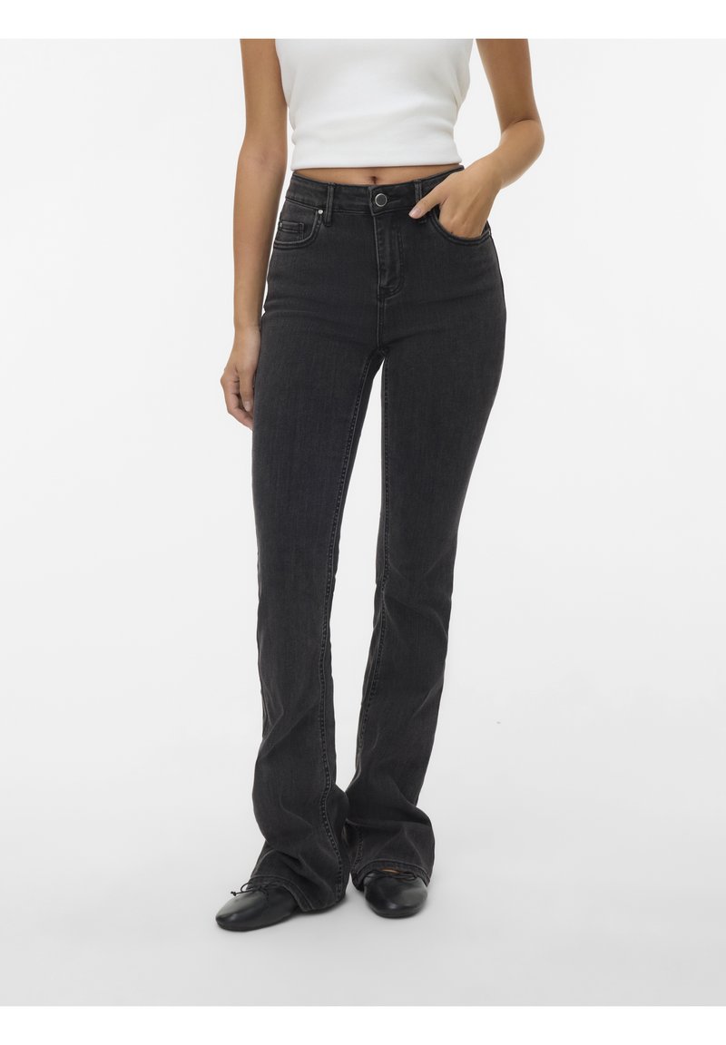 Femme portant un jean noir taille haute, un top court blanc et des chaussures noires, une main dans la poche, sur fond blanc uni.