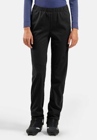 Personne portant un pantalon de sport noir et des chaussures de sport d'hiver noires, se tenant devant un fond blanc uni.