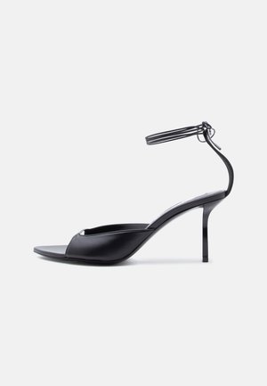 Sandale à talon aiguille en cuir noir avec fine bride à la cheville, bout ouvert et talon modérément haut sur fond blanc.