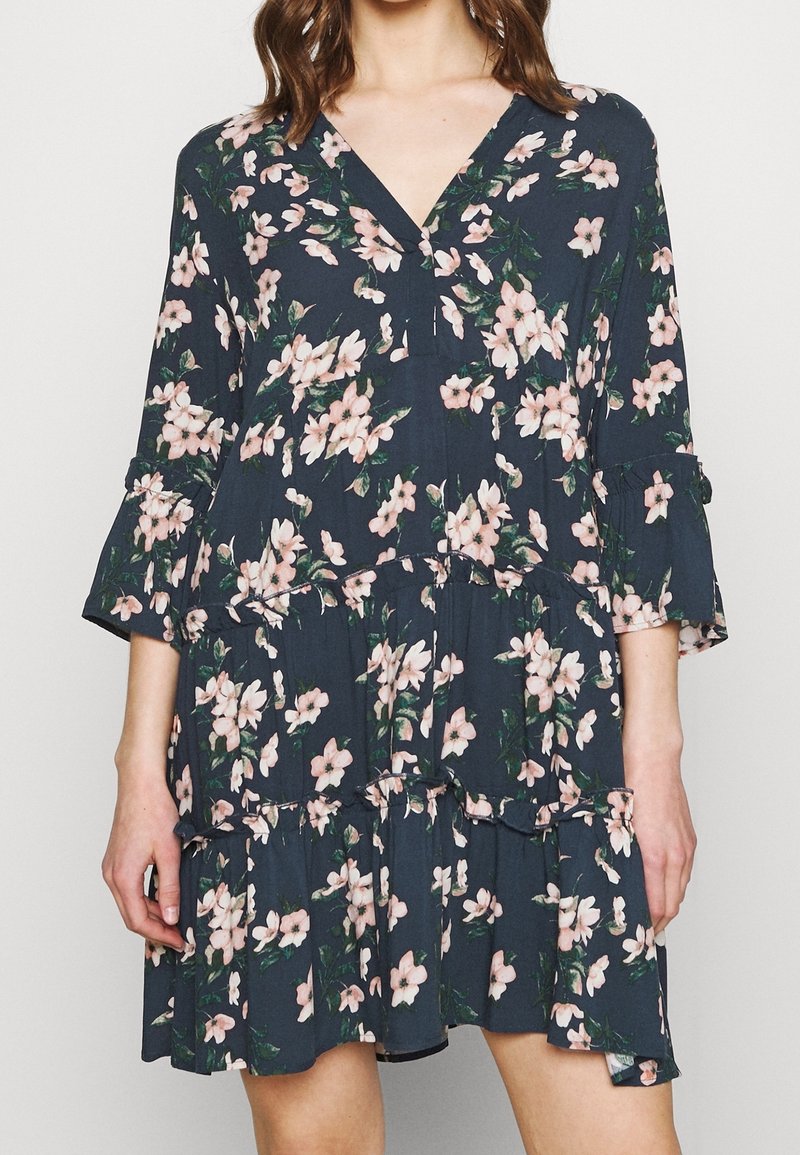 Femme portant une robe fleurie bleu foncé avec des fleurs roses, manches trois-quarts à volants, col en V et jupe à volants.