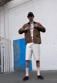 Veste marron avec fermeture à boutons sur un pull blanc, assortie à un short beige et des chaussures marron. Chaussettes unies visibles. Fond industriel.