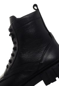 Bota de piel negra hasta el tobillo con un acabado texturizado, parte delantera con cordones y dos botones en el lateral. Cuenta con una suela de goma resistente.