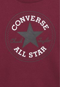 Felpa color marsala con logo circolare che presenta una stella grigia, il testo "CONVERSE" e "ALL STAR" in bianco, e "Chuck Taylor" in corsivo grigio.