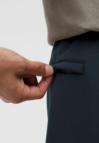 Mørkeblå bukser med en sidelomme. En hånd strækker sig for at åbne lommen. Stoffet ser glat ud med en let strækbar tekstur.