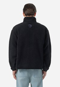 Mężczyzna w czarnej polarowej kurtce z małym okrągłym logo na górnej części pleców, w zestawieniu z jasnoniebieskimi dżinsami, stoi tyłem.