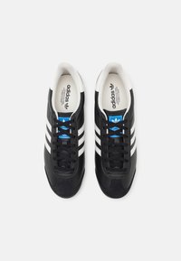 Pár černých tenisek Adidas s bílými pruhy, modrým logem na jazyku a bílým vnitřním podšitím, natočeno zvrchu na jednobarevném pozadí.