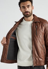 Bruine leren jas met een rits aan de voorkant, gewatteerde schouderdetails en een grijze voering. Voorzien van ritszakken en een zachte textuur.