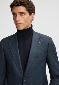 Jonge man met krullend bruin haar, gekleed in een donkerblauwe textuurblazer over een zwarte turtleneck, die iets naar links kijkt op een effen achtergrond.