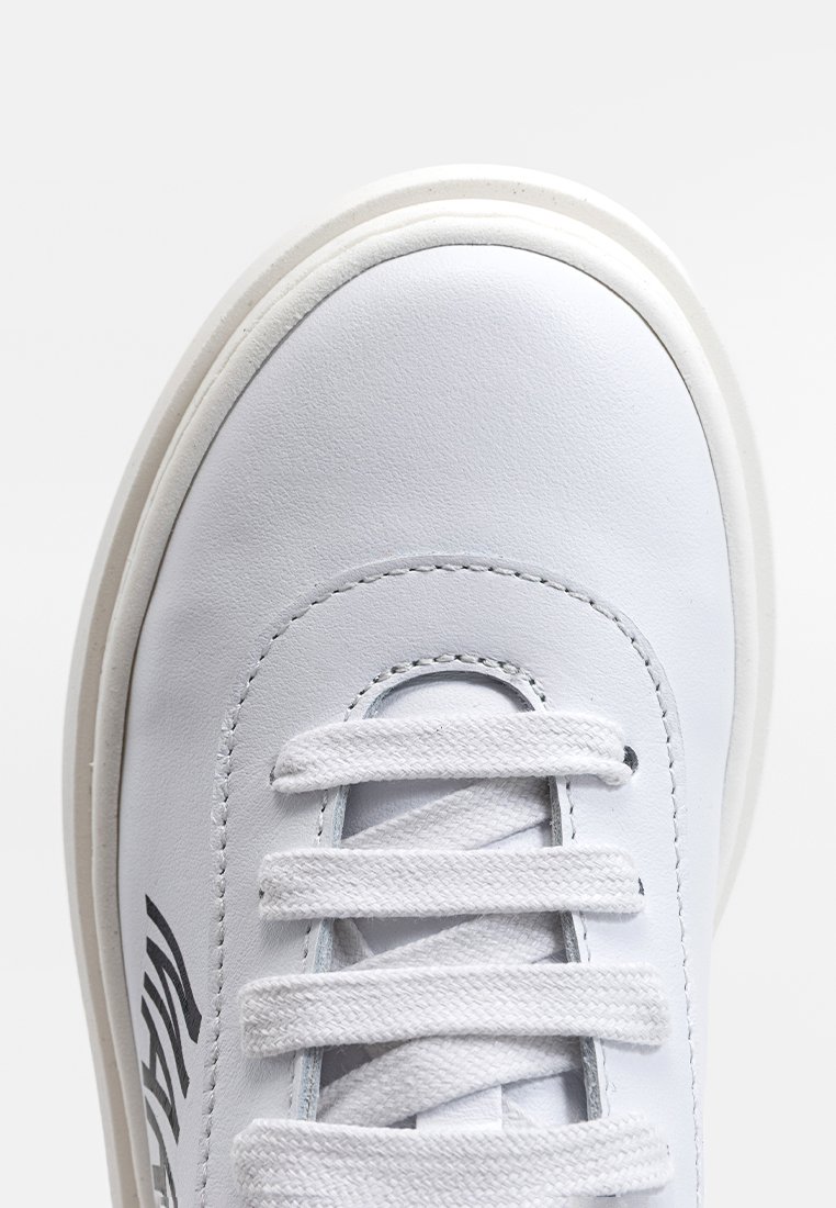 Primo piano della punta di una sneaker bianca in pelle con lacci testurizzati e un logo nero stampato parzialmente sul lato.