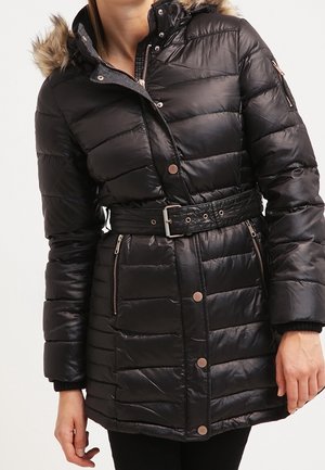 Femme portant un manteau d'hiver matelassé noir ceinturé, avec une capuche bordée de fourrure et des poches zippées, debout les mains détendues.