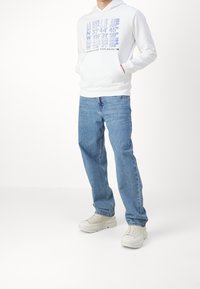Sudadera blanca con capucha y estampado gráfico en azul, combinada con jeans de mezclilla azul claro y zapatillas beige. Ajuste casual, con bolsillos frontales.