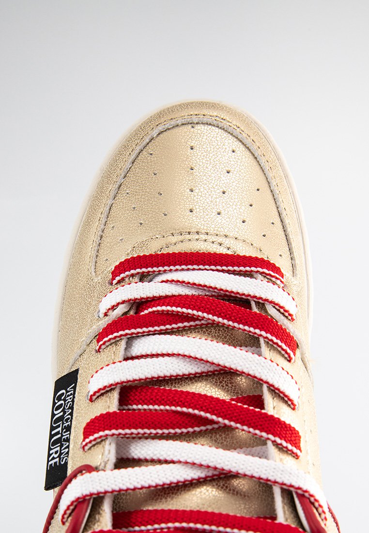 Sneaker in pelle dorata con lacci a strisce rosse e bianche, punta forata e etichetta nera con logo sul lato. Design con suola piatta.