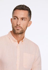 Camisa de botón color melocotón claro, hecha de una tela texturizada, con un bolsillo en el pecho y un cuello con solapa. Sin patrones ni acentos visibles.