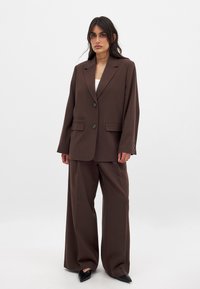Blazer marrón ajustado con dos botones, bolsillos laterales y pantalones amplios a juego; tejido suave con mínimas arrugas, zapatos negros de punta.