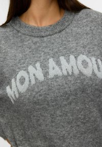 Suéter de punto gris con un cuello redondo y mangas cortas, con el texto "MON AMOUR" en azul claro en la parte frontal.