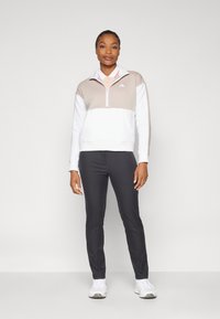 Ελαφρύ πουλόβερ με μισό φερμουάρ σε μπεζ και λευκό, με γιακά και ελαστικές μανσέτες, συνδυασμένο με εφαρμοστά μαύρα παντελόνια και λευκά παπούτσια.