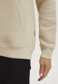 Sudadera beige con acabado texturizado. Incluye un bolsillo canguro, puños acanalados y una pequeña etiqueta de la marca en el dobladillo lateral. Mano visible.