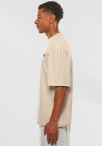Beige oversized t-shirt i bomuld med korte ærmer, har en enkel rund halsudskæring og subtil branding på brystet. Glat tekstur.