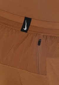 Shorts de sport marron avec une petite étiquette noire Nike et une poche zippée verticale sur le côté droit près de la taille.