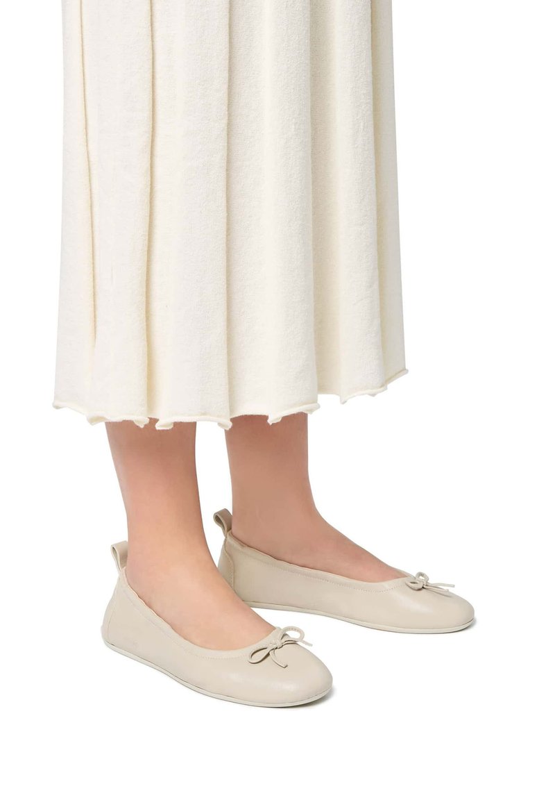 Ballerine in pelle beige con punta arrotondata, dettaglio a grande fiocco sul davanti e suola in gomma flessibile, abbinate a un vestito leggero e strutturato color crema.