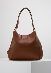 Brown leather handbag with a single wide strap and a magnetic flap closure, displayed on a white pedestal against a plain background.