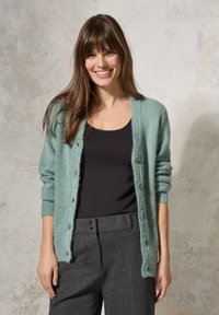 Mintgrön cardigan med en mjuk, fluffig textur, ribbade muddar och sex knappar fram, ihop med en svart topp och grå byxor.