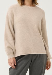 Beiger Strickpullover mit langen, leicht puffigen Ärmeln, gerippten Bündchen und Rundhalsausschnitt. Weicher, strukturierter Stoff, getragen über dunkelgrauen Hosen.