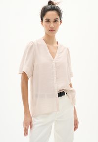 Blusa rosa chiaro con scollo a V, maniche a puff corte e chiusura con bottoni. Realizzata in un materiale morbido e strutturato, abbinata a pantaloni bianchi e una cintura nera.