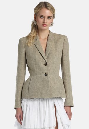 Kvinde iført en tætsiddende beige blazer med to knapper og en hvid plisseret nederdel med frontslids mod en ensfarvet baggrund.