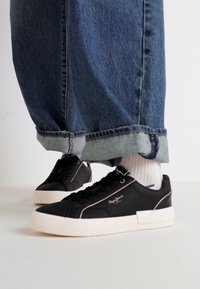 Sneakers neri con suola bianca, tomaia in pelle liscia, dettagli cuciti e logo. Abbinati a jeans in denim blu dalla vestibilità ampia.