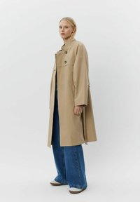Beige trenčkot s vysokým límcem a dvouřadými knoflíky, má dlouhé rukávy, boční kapsy a volný střih, ve spojení s modrými širokými džíny.