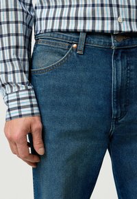 Jean en denim bleu avec une coupe droite, dotée de surpiqûres orange et d'un bouton en cuivre. Associé à une chemise à carreaux bleu, gris et blanc.