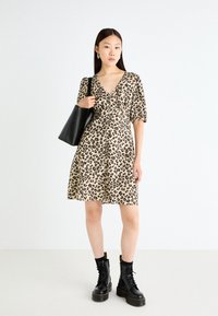 Leopard print dress in beige with a V-neck and short puff sleeves. Model carries a black tote and wears black lace-up boots.