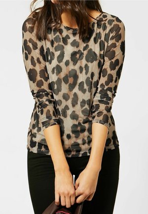Mujer que lleva una camiseta de manga larga con estampado de leopardo marrón y negro, sosteniendo un bolso marrón oscuro delante de pantalones negros.