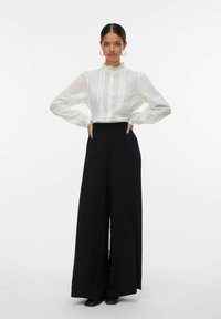 Blouse blanche à manches longues avec col à volants et couture verticale, associée à un pantalon noir à taille haute et jambes larges en tissu doux.