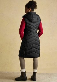 Czarna kamizelka puchowa z kapturem, w wzór chevron, noszona na czerwonym swetrze. W połączeniu z szarymi legginsami w kratkę i brązowymi botkami za kostkę.