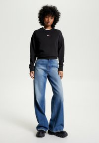 Felpa nera con piccolo logo, abbinata a jeans oversize blu chiaro e scarpe chunky nere con platform, su sfondo neutro.