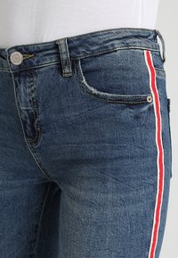 Jeans i mörkblå denim med slitna detaljer. Har en röd sidremsa, fem fickor och en silverfärgad knäppning.