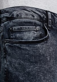 Gros plan sur la poche d'un jean en denim noir délavé à l'acide, avec des coutures visibles, des rivets et une partie d'une chemise grise au-dessus de la taille.