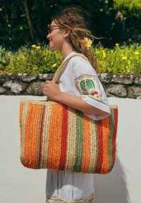 Sac fourre-tout tissé avec des rayures vibrantes en orange, rouge, vert et beige. Comprend des poignées épaisses et une forme spacieuse et structurée.