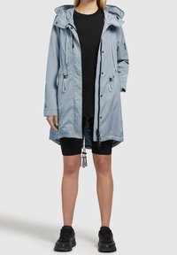 Parka imperméable bleu clair avec capuche à cordon de serrage et taille ajustable, fermeture éclair à l'avant, poches latérales et ourlet réglable, portée par-dessus un haut noir et un short.